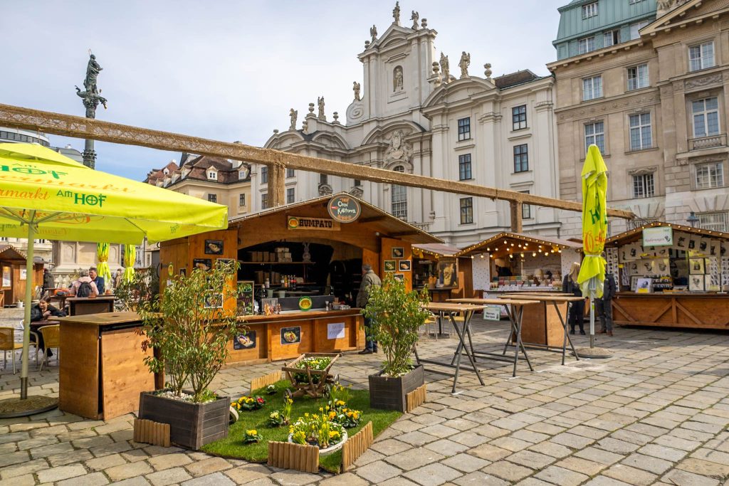 Braune Holzhütten und Frühlingsdeko auf Ostermarkt am Hof in Wien, im Hintergrund eine Kirche.